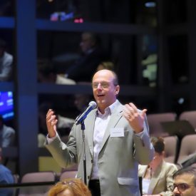 Claudio Mazzali speaks into a standing microphone during the 2026 Corning Materials Innovation Summit, with both hands raised as he gestures. He wears glasses, a light gray blazer, and a white open-collared shirt with a name tag. Audience members sit in tiered seating around him.
