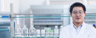 Young man in lab coat, goggles, glass labware in background