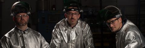 Three men, wearing glass-making protect suits, look into camera