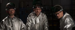 Three men, wearing glass-making protect suits, look into camera