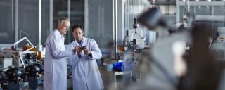 Men in lab coats confer over component in manufacturing setting