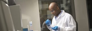 Mann, der einen weißen Laborkittel und eine Schutzbrille trägt, im Hintergrund ein Labor