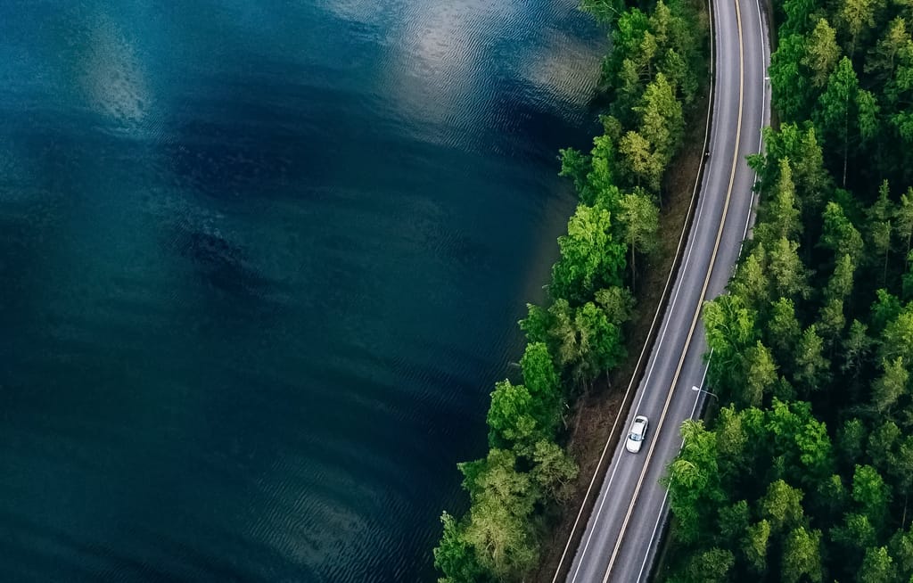 Aerial view of a forested road curving alongside a deep blue lake, with a single car on the highway