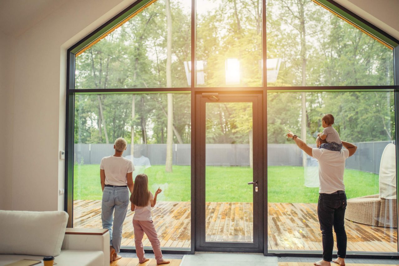 A family stands at a huge window looking out onto the garden while the sun shines back at them