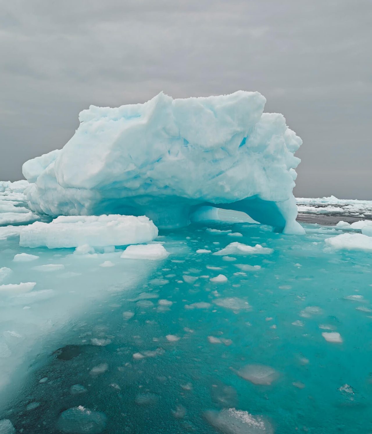 Large chunk of ice floating freely amidst a blue sea. Grey dark sky overhead.