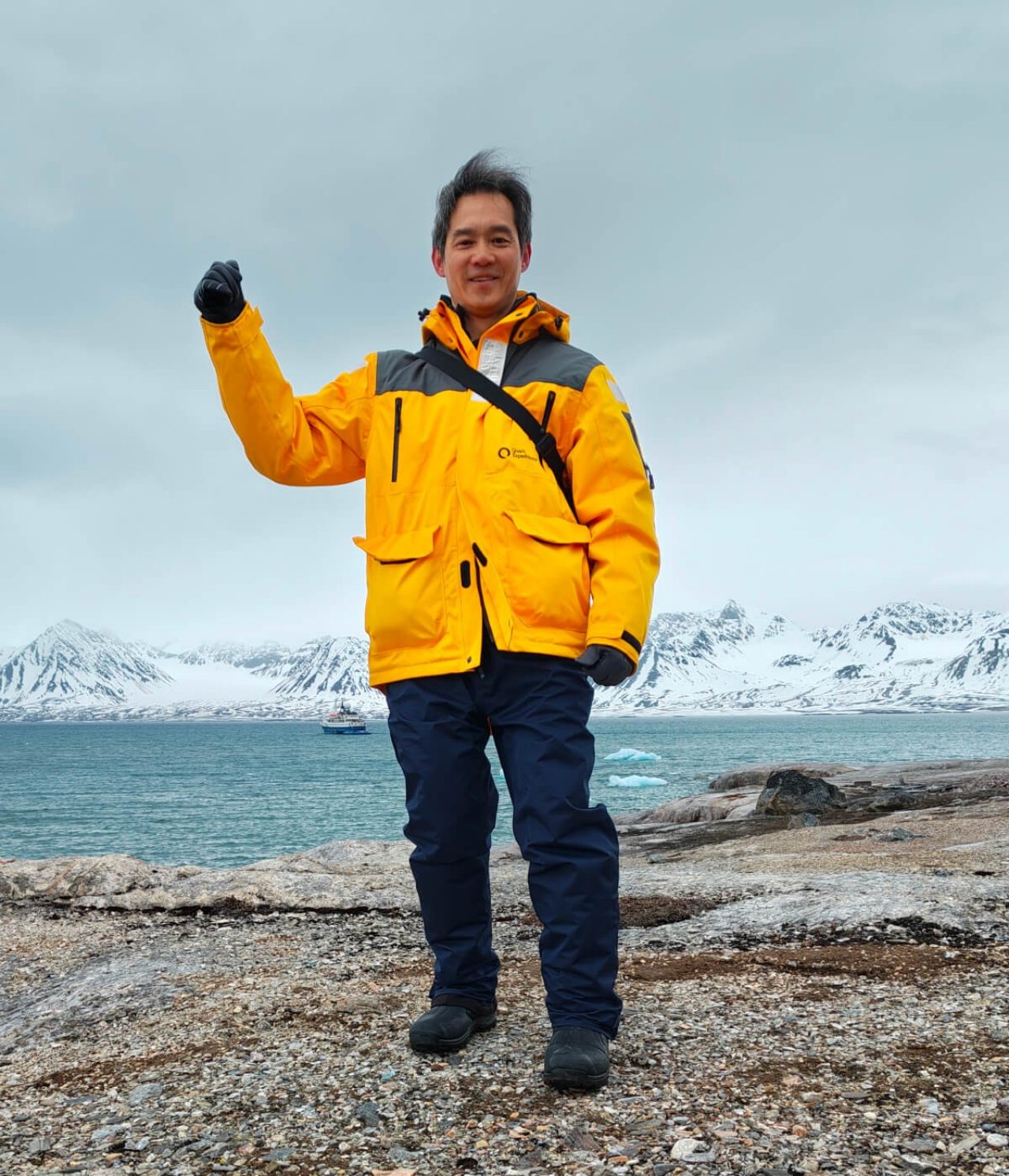 Po Ki in a warm yellow waterproof jacket on a mountain side waving back at the camera