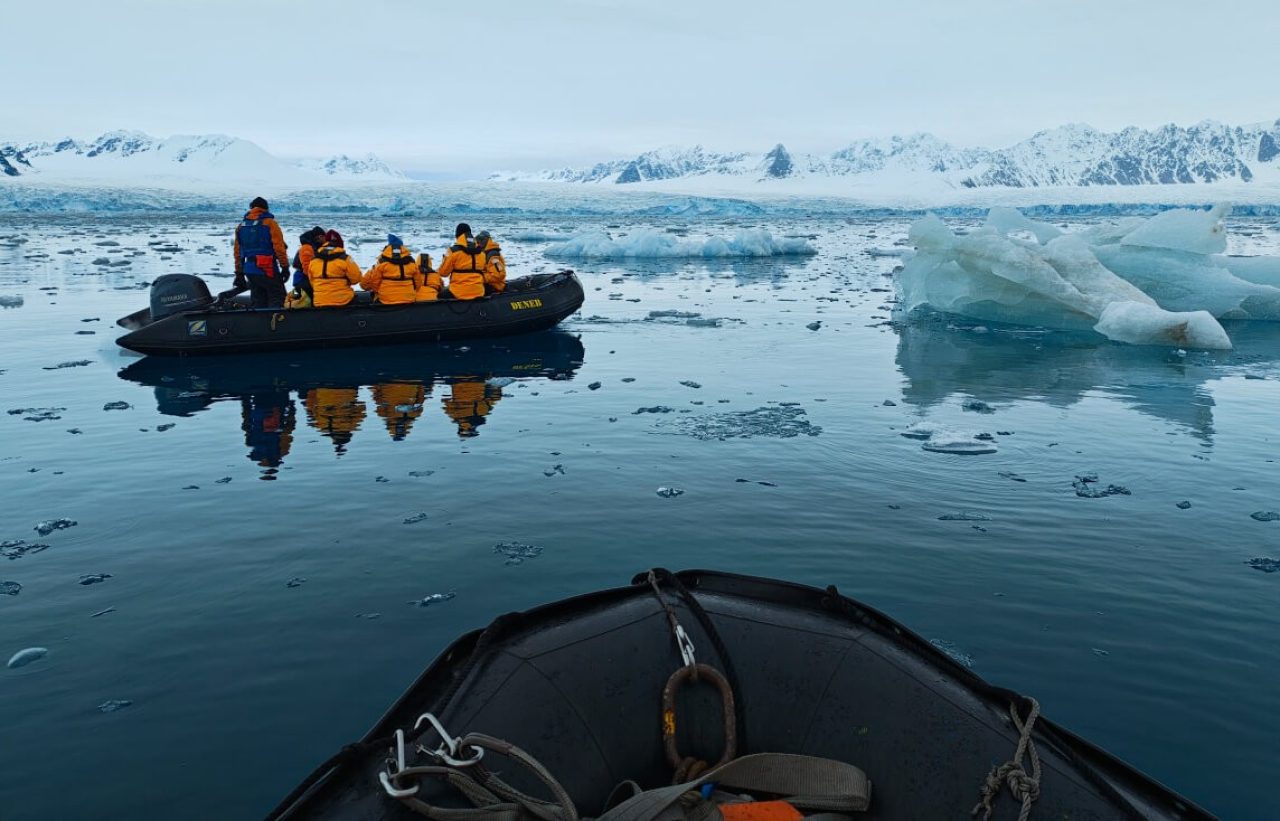 Picture from inside boat looking rib boat with four passengers in orange jackets. Small chunks of ice in water are dotted around the landscape.