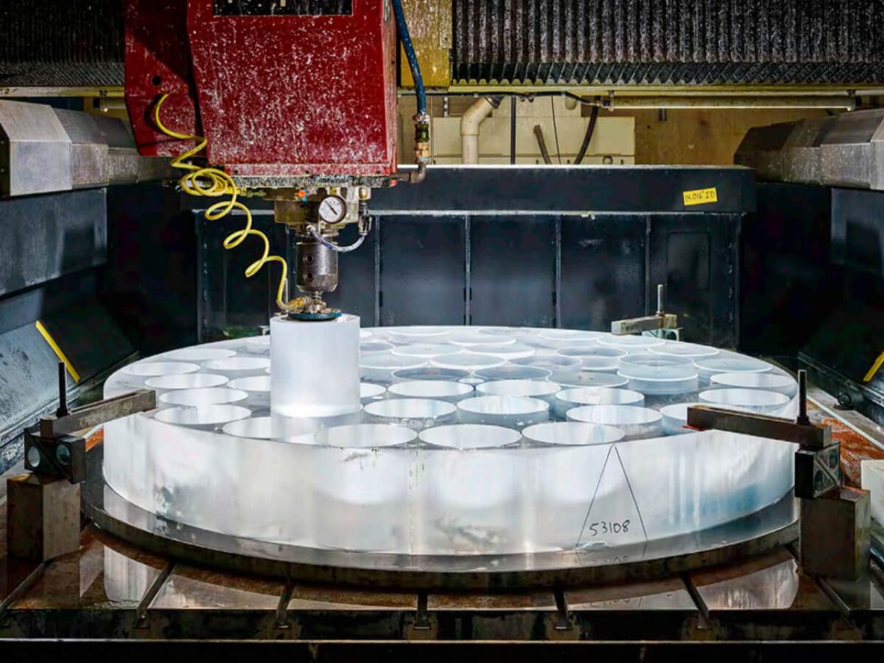 A robotic arm extracts a cylindricle glass core that has been cut from the boule by a machine. The boule is cylindrical opaque glass that is wider than it is tall with many cores already cut out of it.