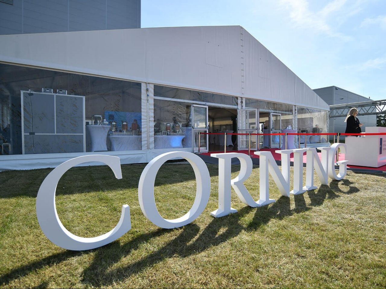 The Corning logo cut out in white foam stands on lawn in front of an events marquee on a bright sunny day. A red carpet is laid out at the entrance.