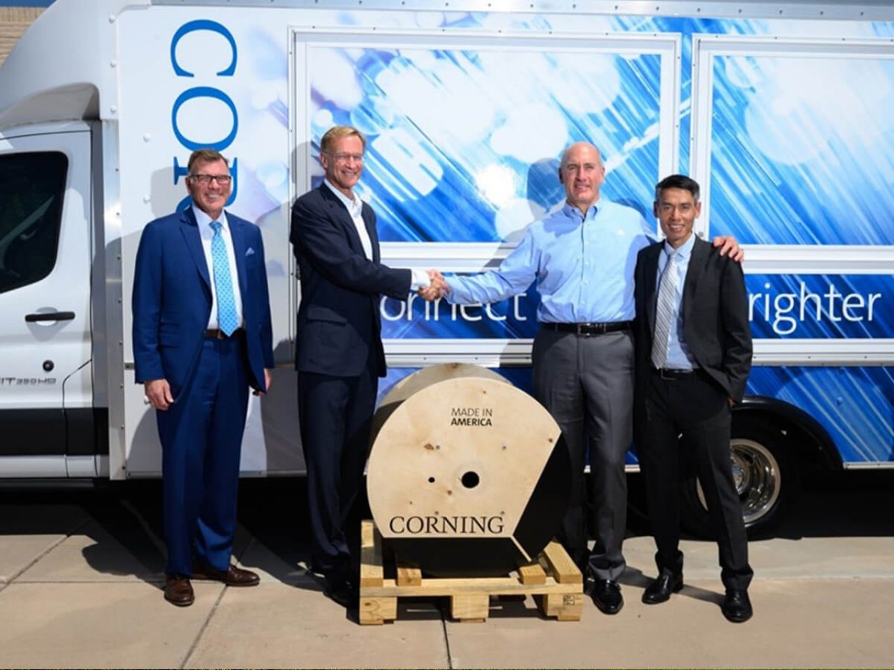 Wendell and Mike shake hands while accompanied by two colleagues. They stand in front of a truck featuring blue Corning livery, with a cable reel on the ground.