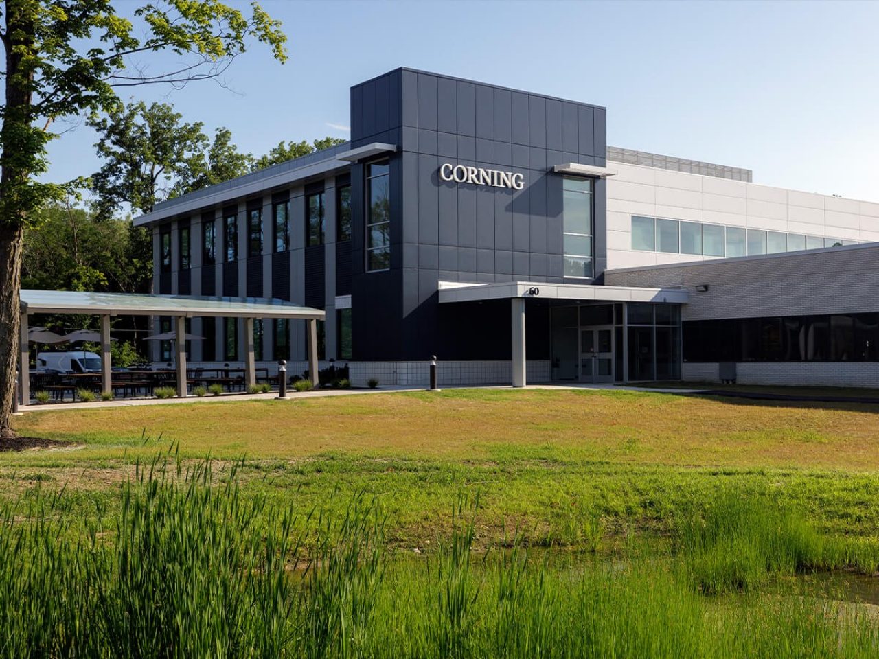 A modern dark and light grey Corning manufacturing facility stands behind green landscape with a small pond in the foreground.