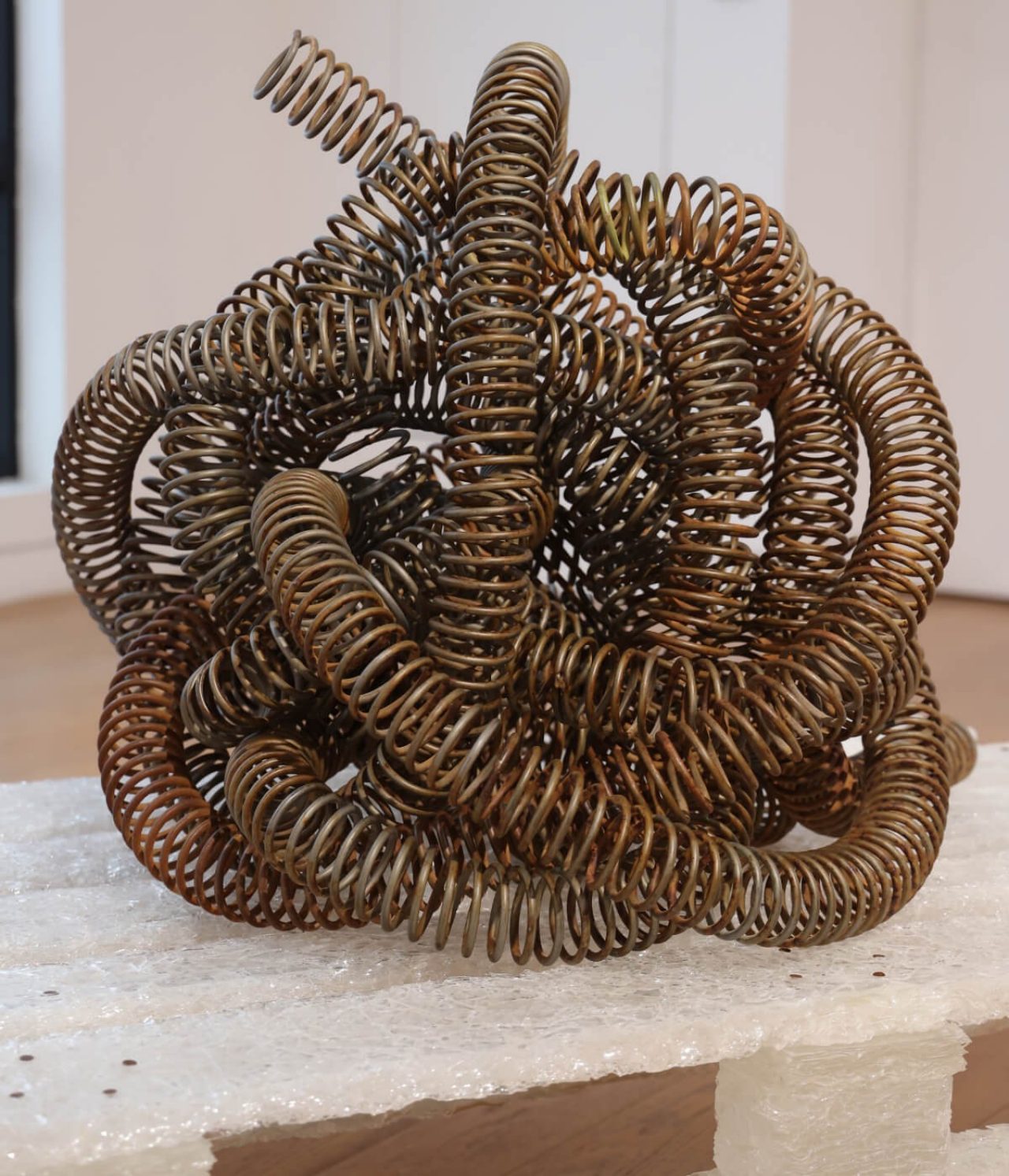 The piece is a metallic coiled mass of furnace wire that sits atop a plinth that looks like a pallet, but made with compressed glass.