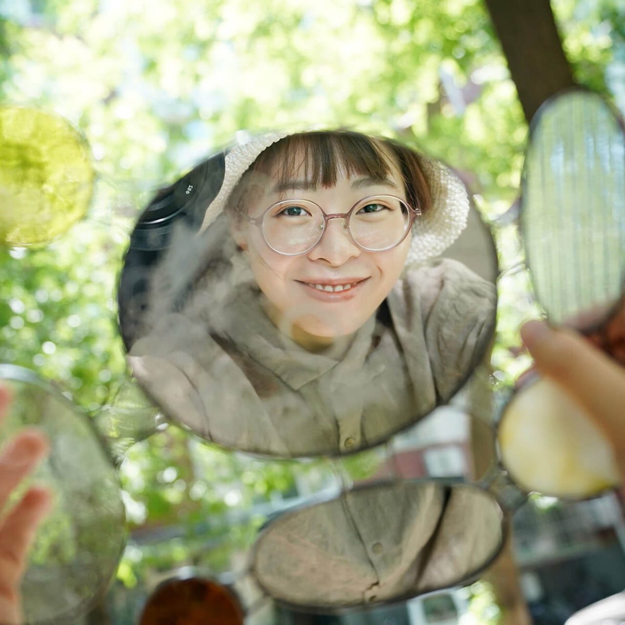 Meng Du wearing glasses, a beige smock and a hat smiles through the reflection of a rounded mirror in a garden setting