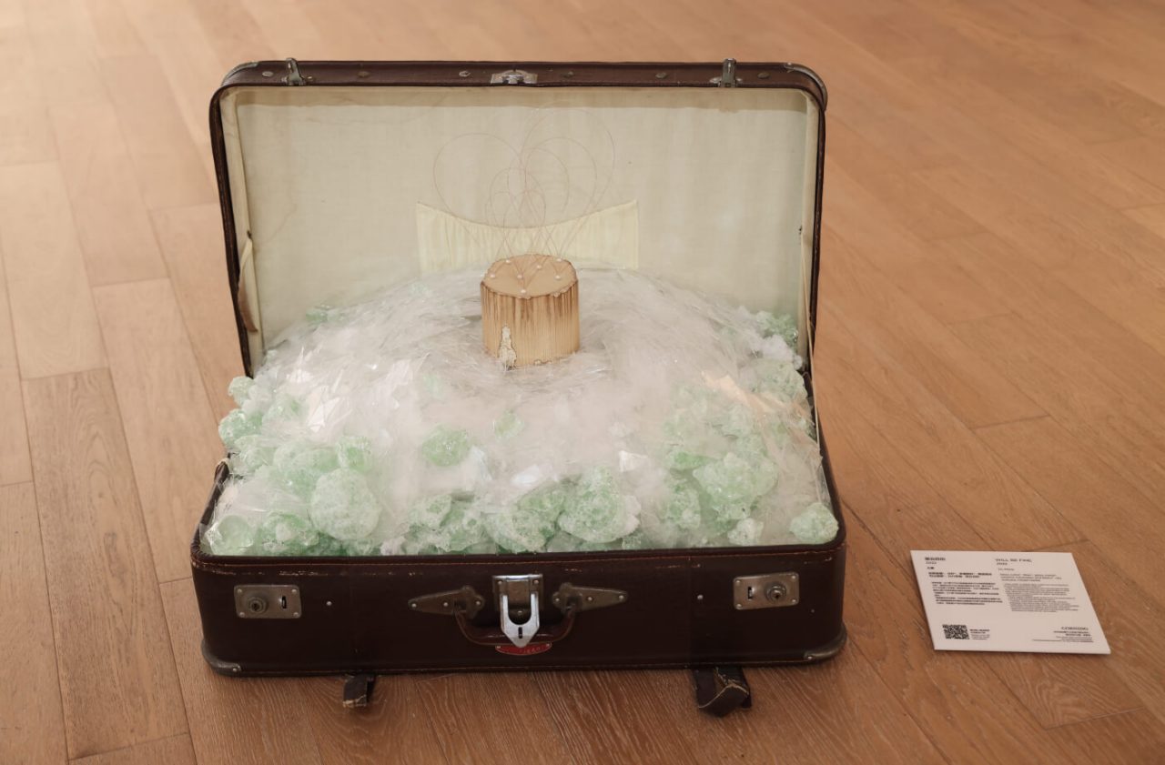 The brown suitcase art piece sits open on the wooden gallery floor. Inside is a crystallized glass-like material.