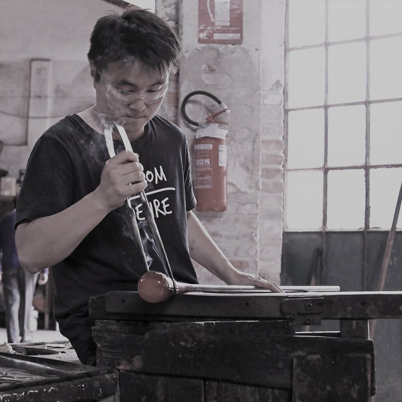 Ji Huang–wearing a black shirt–sits while working on a piece still smoking from heat