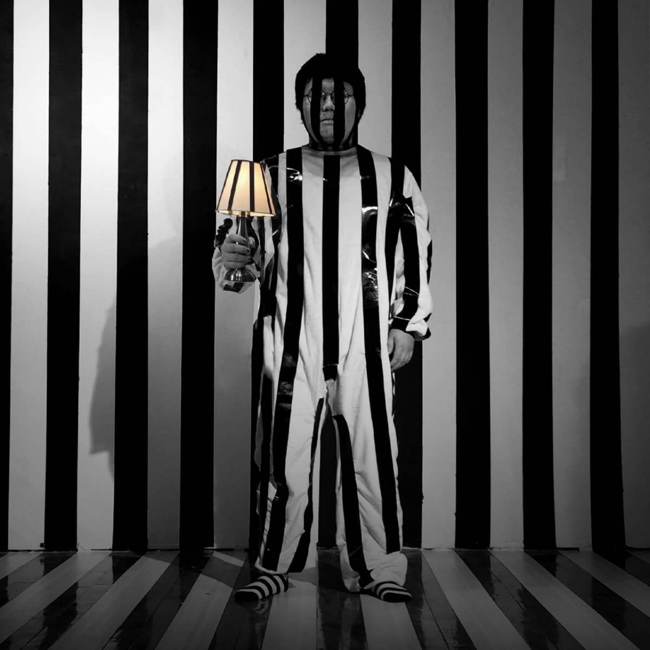 Songlin Li stands with a lamp and shade in his hand against a black and white vertically striped backdrop. The lamp and suit he wears is also striped.